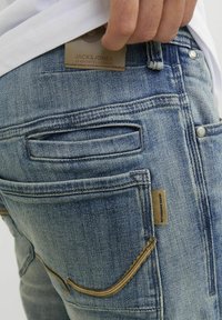 Jeans de mezclilla azul con un acabado desgastado, que presentan múltiples bolsillos, un parche de cuero con el logo y detalles de costura en contraste.