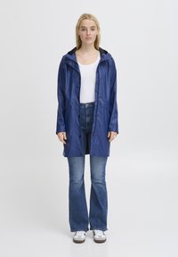 Impermeabile blu con cappuccio, bottoni a pressione e tasche laterali, indossato sopra una maglia bianca e jeans a campana blu. Tessuto morbido.