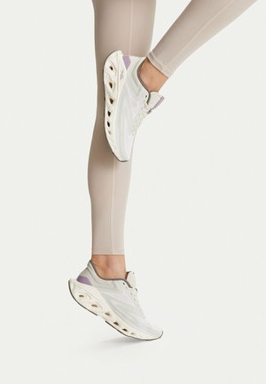 Piernas con leggings beige que llevan zapatillas blancas y lavanda, un pie levantado tocando la pantorrilla opuesta sobre un fondo liso.