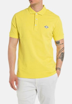 Polo de algodón amarillo con mangas cortas, tapeta de tres botones y un pequeño logo bordado en el pecho. Lleva pantalones blancos.