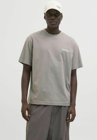 Pelēks kokvilnas T-krekls ar īsām piedurknēm un uzrakstu "UNIVERSAL" baltā krāsā. Apvienots ar brīviem pelēkiem šortiem un baltu cepuri.