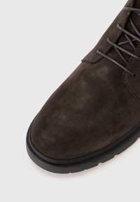 Chaussure en daim marron avec une texture lisse, bout arrondi et semelle en caoutchouc noir. Équipée de lacets et sans motifs ou accents visibles.