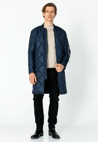 Navy gesteppter langer Mantel mit Stehkragen, Reißverschluss vorne und Seitentaschen; kombiniert mit einem hellen Hemd und schwarzen Hosen. Glatte Textur.