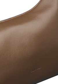 Brun læder skuldertaske med en glat overflade, buet form og præget "Calvin Klein" logo nær den nederste kant.