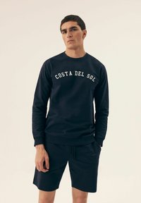 Granatowy sweatshirts z napisem "COSTA DEL SOL" w kolorze białym. Posiada okrągły dekolt i długie rękawy. Nosi się go z pasującymi granatowymi szortami.