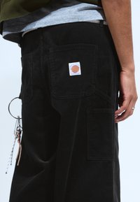 Person mit schwarzen Cordhosen, die einen Patch auf der Gesäßtasche haben, und Schlüsselanhänger, die von der Schlaufe der Seitentasche hängen.