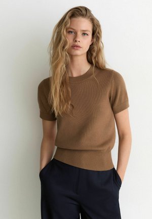 Mujer con cabello largo y rubio que lleva un top de punto marrón de manga corta y pantalones oscuros, de pie contra una pared lisa y clara con las manos en los bolsillos.