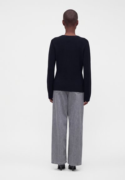 Personne portant un pull noir à manches longues et un jean gris ample, debout dos à la caméra sur un fond uni.