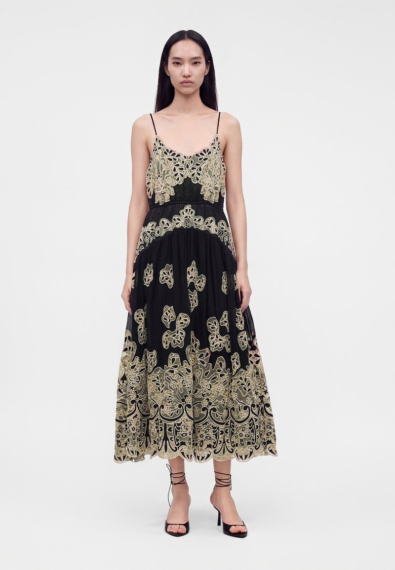 Woman wearing a black midi dress with beige floral embroidery and thin straps, paired with black strappy high-heeled sandals.