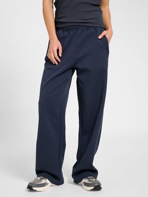 Personne portant un pantalon large bleu marine avec taille élastique et baskets grises, debout avec une main dans la poche devant un fond uni.