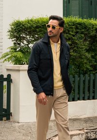 Chaqueta de marino con cierre de cremallera, polo beige y pantalones color marrón claro. Gafas de sol amarillas añaden contraste. Texturas suaves y ajuste a medida. Follaje verde en el fondo.