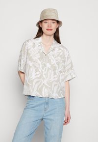 Camisa curta e cortada em bege claro com padrão floral branco. Frente com botões e ajuste relaxado. Usada com jeans azul claros.