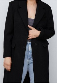 Cappotto in misto lana nero con design strutturato, chiusura a singolo bottone e rever a punta. Indossato sopra una canottiera grigia e jeans blu.