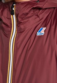 Giacca K-Way color amaranto con zip a righe bianche, arancioni, blu navy e gialle, mostrante la parte anteriore e il logo sulla zona del petto.