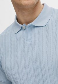 Svetlo modra polo majica z teksturiranim pletenim vzorcem in navpičnimi črtami, dvema gumboma na vratu in klasičnim ovratnikom.