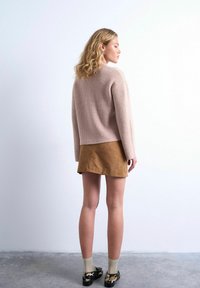 Femme aux cheveux blonds ondulés portant un pull en maille beige, une mini-jupe en daim marron, des chaussettes crème et des chaussures imprimé léopard, debout contre un mur blanc.