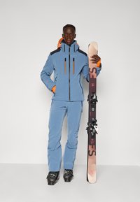 Ski apģērbs gaiši zilos toņos, ar melniem un oranžiem akcentiem, satur zipu jaku, saskaņotas bikses un melnus slēpošanas zābakus, kas tur slēpi.