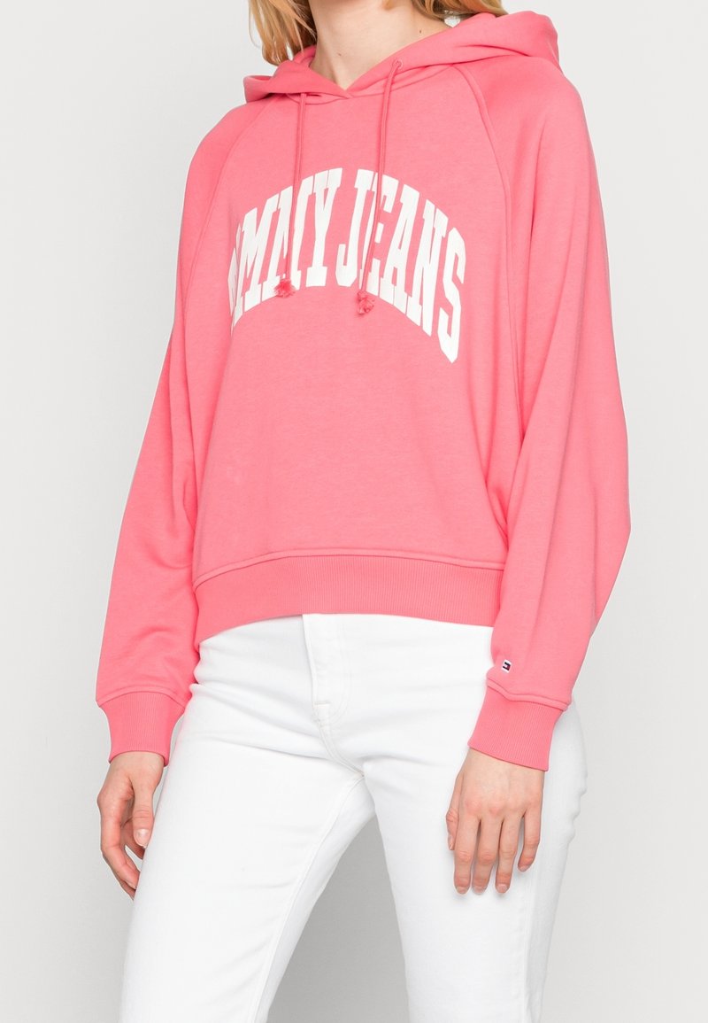 Sweat à capuche rose en tissu doux, avec une poche kangourou à l'avant, une capuche avec cordons de serrage, et le logo "TOMMY JEANS" en lettres blanches en gras.