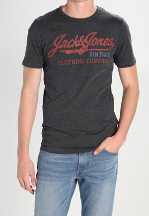 Mężczyzna noszący ciemnoszarą koszulkę Jack & Jones Vintage Clothing Company i jasnoniebieskie dżinsy, z jedną ręką w kieszeni.
