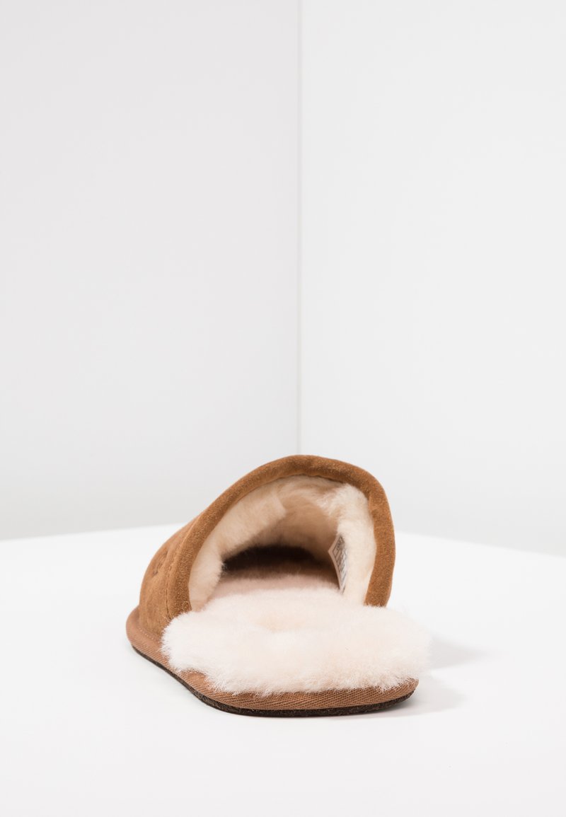 Braune Wildleder-Hausschuhe zum Hineinschlüpfen mit einem weichen, cremefarbenen Fellfutter, abgerundeter offener Zehenpartie und flacher Gummisohle. Schlichtes Design, ohne Verzierungen.