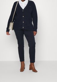 Navy knit cardigan with a v-neck, button front, and long sleeves, paired with dark trousers and brown heeled lace-up ankle boots.