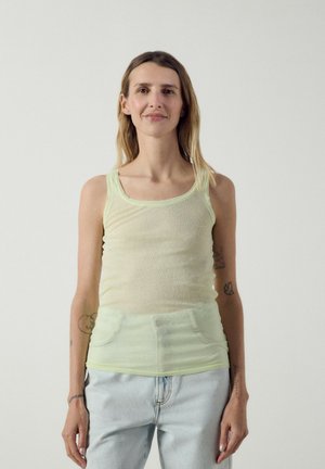 Frau mit Tattoos, die ein hellgrünes ärmelloses Top und hellblaue Jeans trägt, vor einem schlichten hellen Hintergrund stehend.