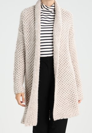 Henkilö pukeutuneena beigeen, ylipaksuun neulottuun isoon neuletakkiin mustavalkoraidallisen poolopaidan ja mustien housujen päällä.