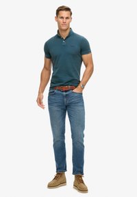 Polo de color verde azulado oscuro con cuello y dos botones, combinado con jeans azul claro y botas trenzadas color tostado. Dise�ño casual y ajustado.