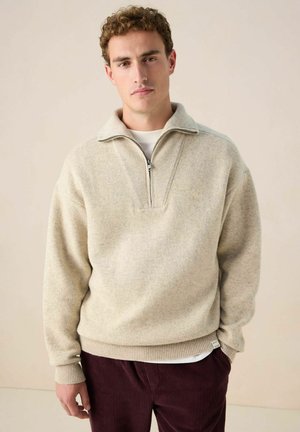 Man with curly hair wearing a beige quarter-zip sweater over a white shirt and dark red corduroy pants, standing against a plain background.