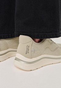 Bežīgas tekstila sporta apavi ar mīkstu zoli, kurām ir struktūrēta virsma un sudraba akcenti uz papēža, novietoti blakus tumšiem biksēm.