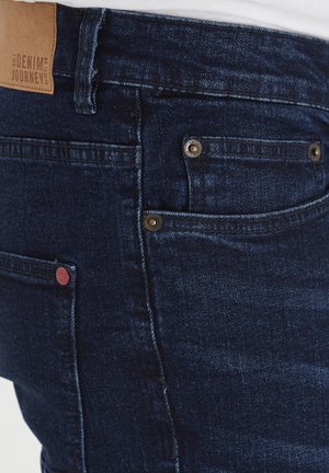 Tumedav sinised teksad, millel on sile tekstuur, messingist tarvikud taskutel ja pruun nahast silt "The Denim Journey."