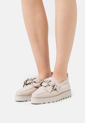Loafers plateformes beige clair avec des détails en chaîne argentée et un nœud en tissu, portés pieds nus sur fond blanc.