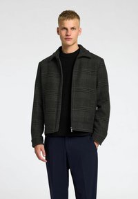 Veste à carreaux gris foncé en tissu texturé, avec un col, une fermeture éclair et une silhouette structurée, portée sur un pull en tricot noir.