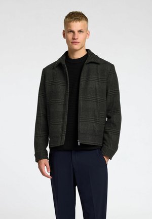 Veste à carreaux gris foncé en tissu texturé, avec un col, une fermeture éclair et une silhouette structurée, portée sur un pull en tricot noir.