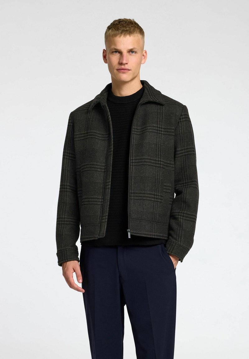 Veste à carreaux gris foncé en tissu texturé, avec un col, une fermeture éclair et une silhouette structurée, portée sur un pull en tricot noir.