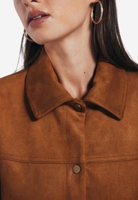 Brun mockakjakett med nedfällbar krage, med knappknäppning och synliga sömnadsdetaljer. Strukturerad, mjuk yta.