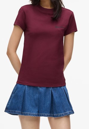 Kvinde iført en kortærmet vinrød t-shirt og et plisseret blåt denim miniskørt, stående med hænderne bag ryggen.