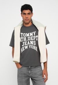 Pelēks kokvilnas T-kreklis ar lieliem baltiem burtiem, kas lasa "TOMMY ATH. DEPT. JEANS NEW YORK," apvienots ar krēmkrāsas un piparmētru flīsa puloveri, kas uzkārts pār pleciem.