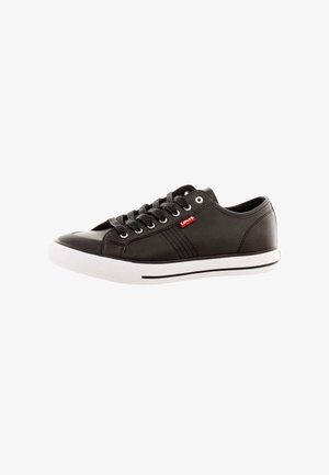 Baskets en cuir noir avec un bout rond, dotées de lacets gris, d'une semelle en caoutchouc blanche et d'une étiquette logo rouge. Texture lisse avec des accents de design minimalistes.