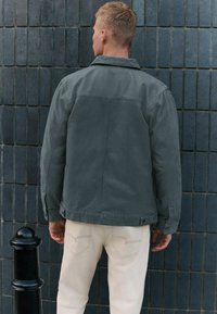 Veste en denim grise avec un col, vue de dos. La silhouette porte un pantalon de couleur claire. Une texture subtile et des détails de couture sont visibles.