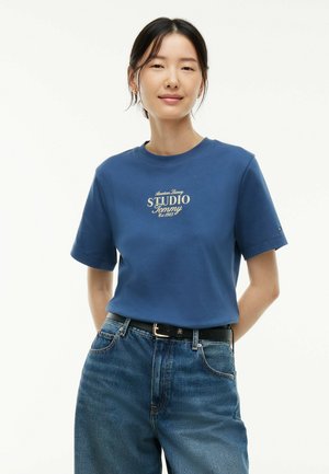 Femme aux cheveux noirs attachés, portant un t-shirt bleu Studio Tommy et un jean bleu taille haute avec une ceinture noire, debout devant un fond blanc.