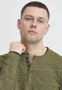Tamsiai žalia ilgomis rankovėmis marškiniai su tekstūruotu audiniu, trijų mygtukų apykakle ir kontrastingais juodais apykaklės bei rankovių kraštais.