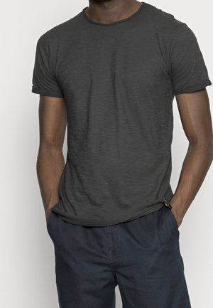 Mann in einem enganliegenden schwarzen T-Shirt mit kurzen Ärmeln und einer navyblauen Hose, mit Händen in den Taschen vor einem schlichten Hintergrund.