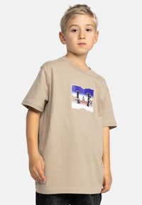 Beige bomull T-shirt med en stor flerfärgad DC-logotyp på framsidan. Korta ärmar och avslappnad passform, visad på en ung modell.