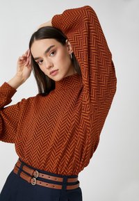 Pomarańczowy sweter w wzór w warkocz, z wysokim dekoltem i wyolbrzymionymi rękawami, zestawiony z ciemnym, paskiem w talii.