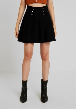 Czarna plisowana spódnica z marszczonymi akcentami i guzikami przypominającymi perły na pasku, wykonana z teksturowanej tkaniny. Noszona z czarnymi botkami za kostkę.