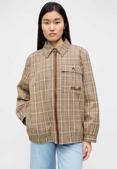 Τζάκετ ταρτάν σε ανοιχτό κρεμ χρώμα με καφέ φερμουάρ, τσέπη στο στήθος με κουμπί και μανίκια με μανσέτες. Το υλικό φαίνεται ελαφρύ με λεία υφή.