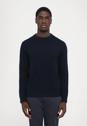Jeune homme avec les cheveux courts portant un pull uni bleu marine et un pantalon sombre, se tenant devant un fond gris clair.
