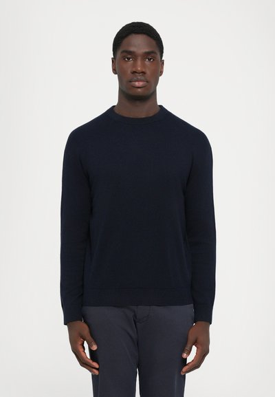 Ung mand med kort hår iført en ensfarvet mørkeblå sweater og mørke bukser, stående foran en lys grå baggrund.