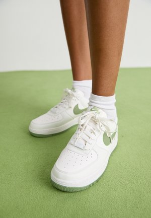 Osoba nosząca białe buty Nike Air z jasnokołowym znakiem i białe skarpetki stojąca na zielonym dywanie.